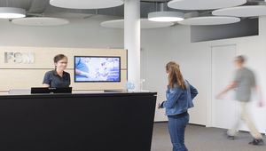 Bildbeschreibung: Empfangsbereich der FSM AG. Eine Mitarbeiterin steht hinter dem Empfangstresen und spricht mit einer Besucherin. Im Hintergrund ist ein Bildschirm mit einer Präsentation sowie eine helle Wand sichtbar. Ein weiterer Mensch bewegt sich in Richtung des Durchgangs auf der rechten Seite.